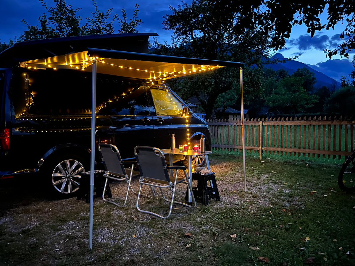Camping van standing on a camp ground in the evening with a table and chairs infront of it and fairy lights glistening